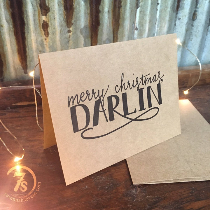 The Darlin' Christmas Card by Savannah Sevens western life{&}style features “Merry Christmas, Darlin’” in black script on kraft paper. Displayed on wood with string lights, it comes with an envelope and a circular 7s logo for a charming holiday touch.