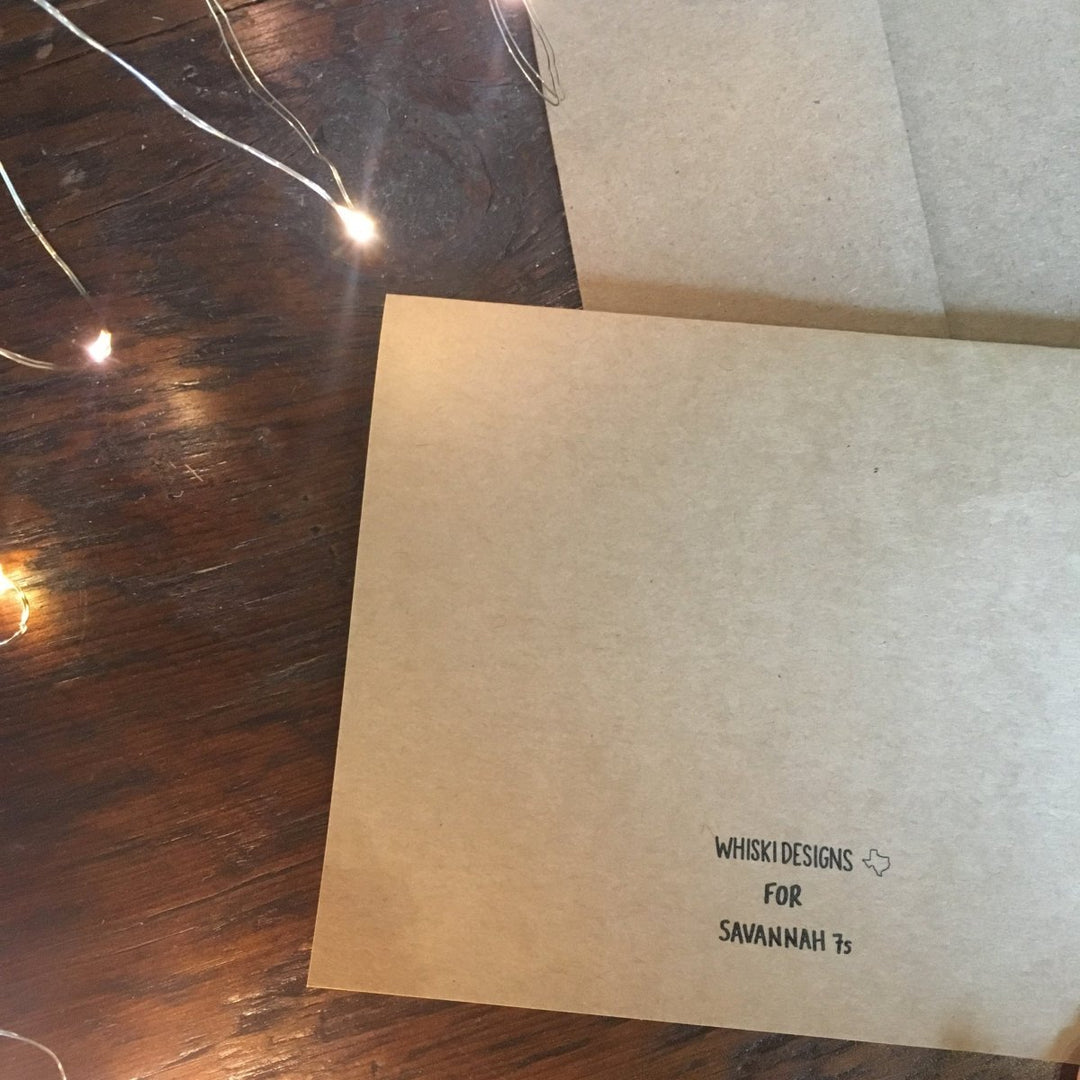 A brown envelope on a wooden surface with string lights nearby features a stamp reading Savannah Sevens western life{&}style—perfect for mailing your Darlin' Christmas Card this season.