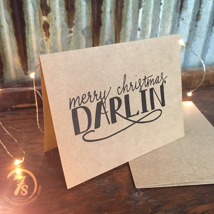 The Darlin' Christmas Card by Savannah Sevens western life{&}style features “Merry Christmas, Darlin’” in black script on kraft paper. Displayed on wood with string lights, it comes with an envelope and a circular 7s logo for a charming holiday touch.