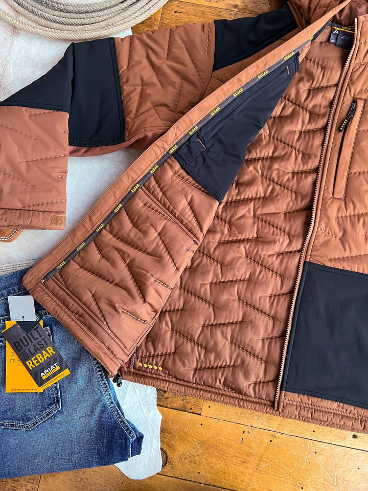 A Davenport Ariat Men's Insulated Jacket by Savannah Sevens western life{&}style is unzipped to show its lining, lying next to blue jeans with a Built for Rebar tag on a wooden floor, with a coil of rope in the background.