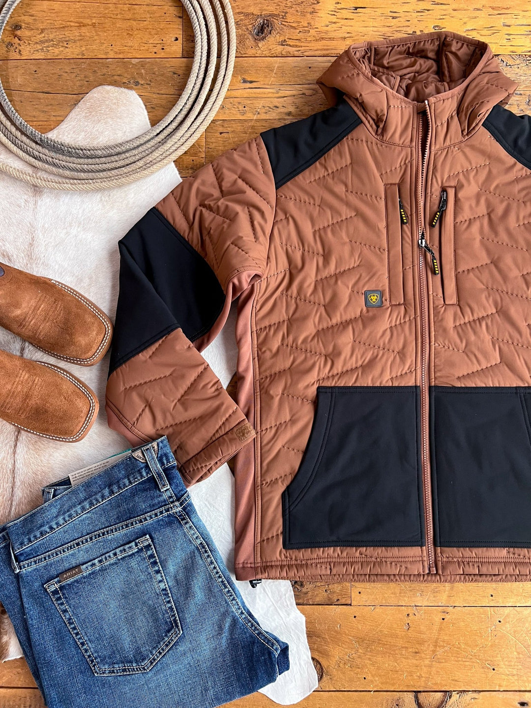 The Davenport Ariat Men's Insulated Jacket by Savannah Sevens western life{&}style, in brown and black, is displayed with a coiled rope, brown slip-on shoes, blue jeans, and a white textured mat on a wooden floor.