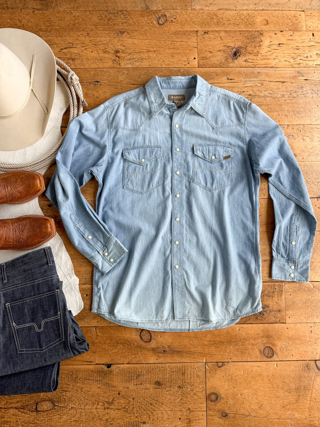 The Davidson Ariat Men’s Denim Pearl Snap shirt from Savannah Sevens western life{&}style is laid flat on a wooden floor. It’s light blue with pearl snaps and displayed alongside brown boots, dark jeans, a white cowboy hat, and a coiled rope.