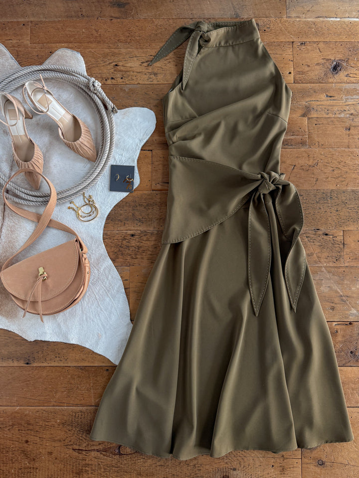 The Decatur Olive Halter Neck Dress by Savannah Sevens western life{&}style is laid out on a wooden floor, paired with beige heels, a tan crossbody bag, gold hoop earrings, and a light-colored cloth.