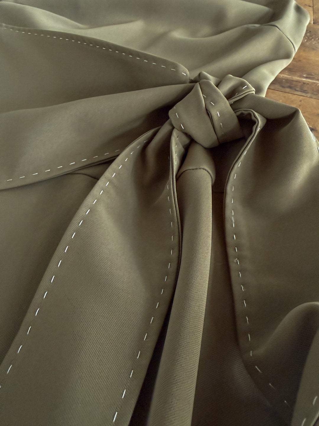 A close-up view of olive green fabric with white basting stitches, gathered and tied in a knot, laid on a wooden surface—featured in the Decatur Olive Halter Neck Dress by Savannah Sevens western life{&}style. Ideal for designing an adjustable tie dress or full-length dress.