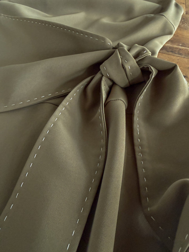 A close-up view of olive green fabric with white basting stitches, gathered and tied in a knot, laid on a wooden surface—featured in the Decatur Olive Halter Neck Dress by Savannah Sevens western life{&}style. Ideal for designing an adjustable tie dress or full-length dress.