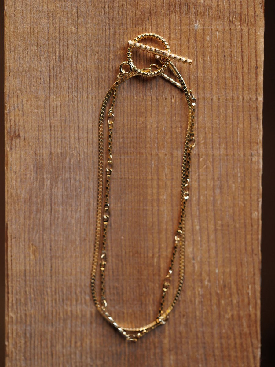 The Declo Necklace by Savannah Sevens western life{&}style, featuring intertwined fine gold chains and a twisted toggle clasp, is displayed on a textured wooden surface for an elegant layered look.
