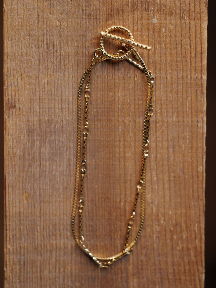 The Declo Necklace by Savannah Sevens western life{&}style, featuring intertwined fine gold chains and a twisted toggle clasp, is displayed on a textured wooden surface for an elegant layered look.