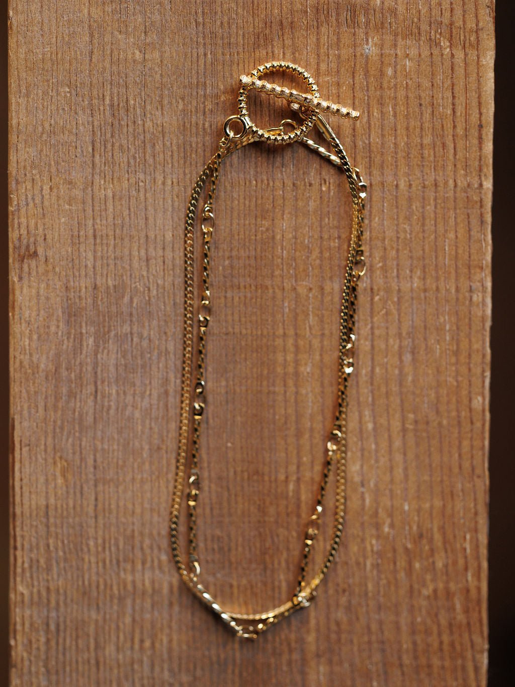 The Declo Necklace by Savannah Sevens western life{&}style, featuring intertwined fine gold chains and a twisted toggle clasp, is displayed on a textured wooden surface for an elegant layered look.