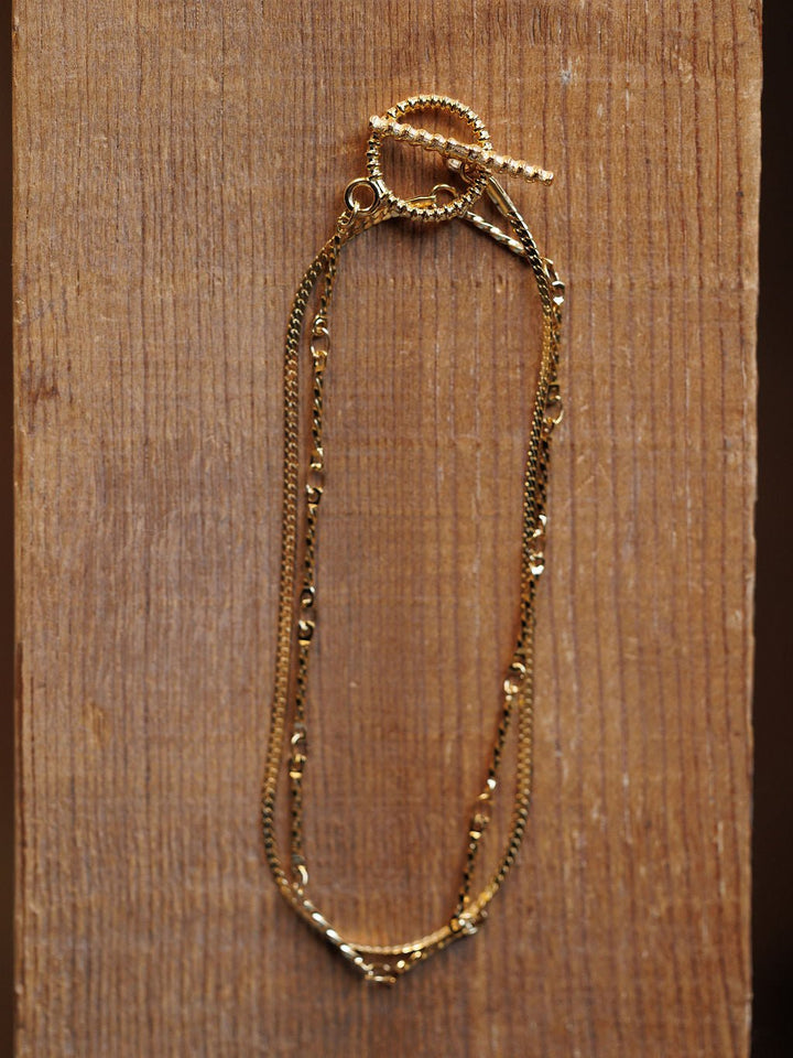 The Declo Necklace by Savannah Sevens western life{&}style, featuring intertwined fine gold chains and a twisted toggle clasp, is displayed on a textured wooden surface for an elegant layered look.