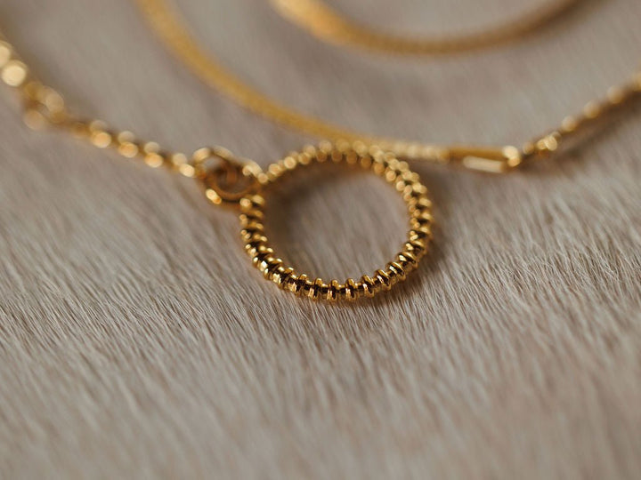 A close-up of the Declo Necklace by Savannah Sevens western life{&}style shows its gold filled stainless steel chain and textured, waterproof circular pendant on a light wooden surface.