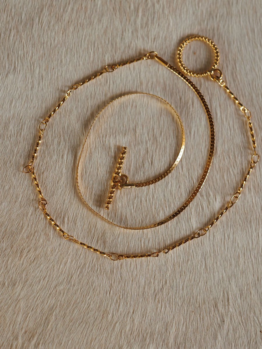 The Declo Necklace by Savannah Sevens western life{&}style, along with two other gold necklaces including a dual strand and a waterproof style, are displayed on a light tan, cowhide surface.