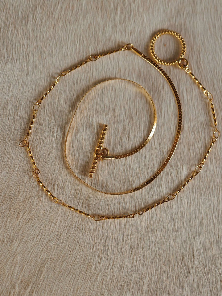 The Declo Necklace by Savannah Sevens western life{&}style, along with two other gold necklaces including a dual strand and a waterproof style, are displayed on a light tan, cowhide surface.