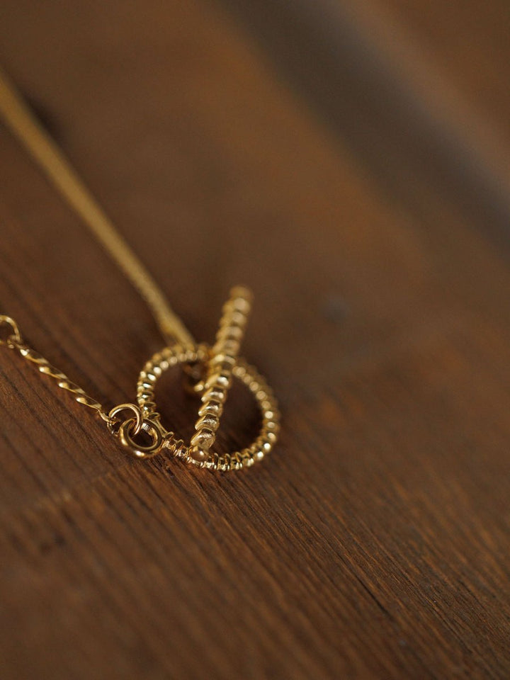 A close-up of the Declo Necklace by Savannah Sevens western life{&}style, featuring a gold filled stainless steel design and beaded toggle clasp, resting on a wooden surface.