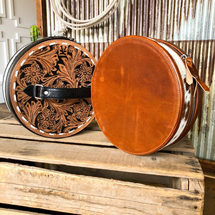 Two round Del Rey Makeup Cases by Savannah Sevens western life{&}style rest on a rustic wooden surface: one features intricate tooled floral leather designs, the other is plain brown with a hair-on-hide accent. Both have handles and zippers.