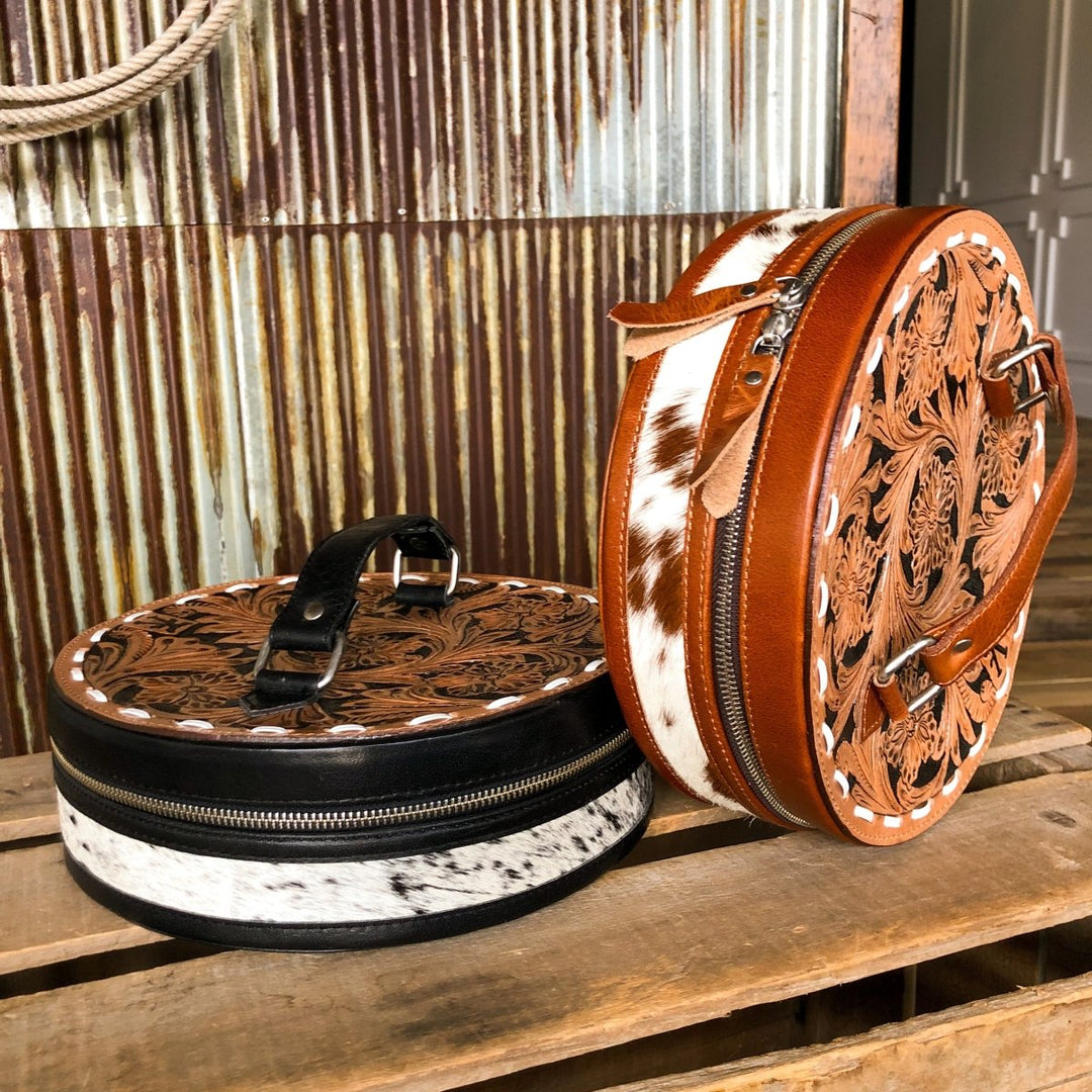 Two Del Rey Makeup Cases by Savannah Sevens western life{&}style—a black and white, and a brown and white tooled leather with floral designs—rest on a wooden crate; one features a shoulder strap.