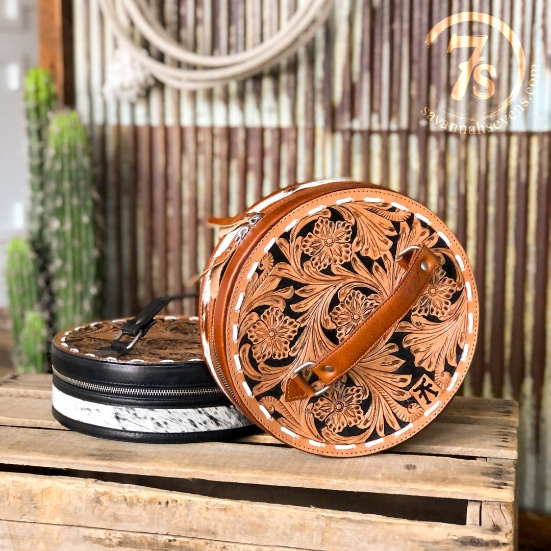 Two round Del Rey Makeup Cases by Savannah Sevens western life{&}style, one tan handmade leather with a handle and one with a dark hair on hide pattern, sit on rustic wood with a cactus and metal wall in the background.