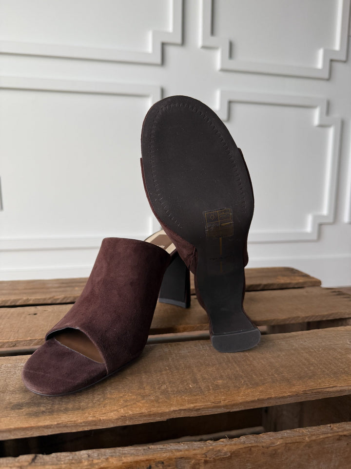 The Delano Mahogany Suede Heel by Savannah Sevens western life{&}style is displayed on a wooden surface, with one shoe standing upright and the other revealing the cushioned footbed and sole. A white panel wall serves as the backdrop.