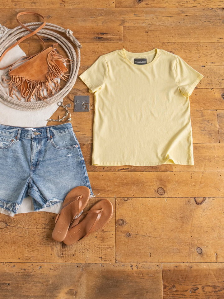 A casual summer outfit on a wooden floor featuring the Denning Butter Tee by Savannah Sevens western life{&}style, distressed denim shorts, tan flip flops, a tan fringed crossbody bag with a rope strap, and earrings.