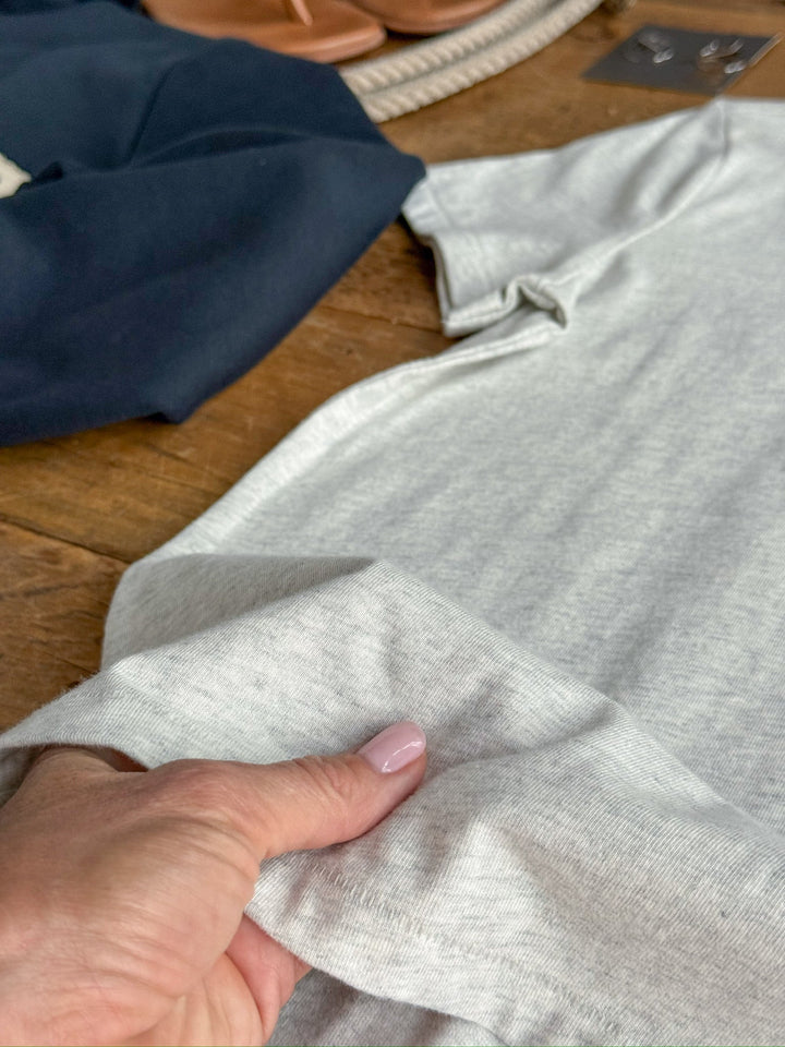 A hand holds the hem of the Savannah Sevens western life{&}style Denning Heather Grey Basic Tee, featuring a semi-relaxed fit, displayed on a wooden surface with a navy garment and tan sandals in the background.