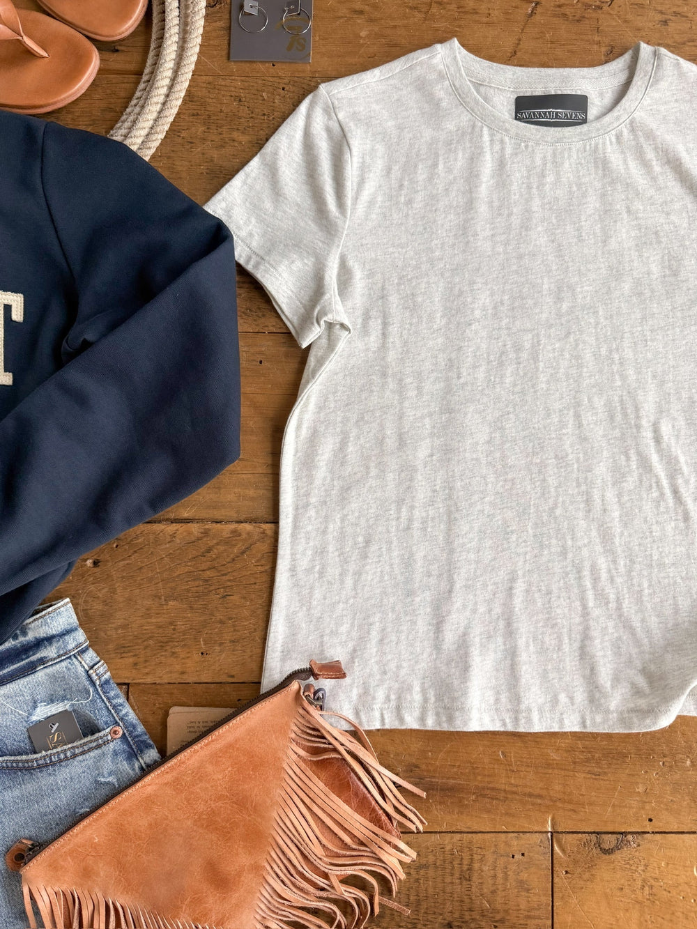 Flat lay of the Denning Heather Grey Basic Tee by Savannah Sevens western life{&}style, paired with blue jeans, a navy sweatshirt, tan sandals, a brown fringe clutch, and rope accessories on a wooden surface. This cotton short sleeve top offers a semi-relaxed fit for effortless style.