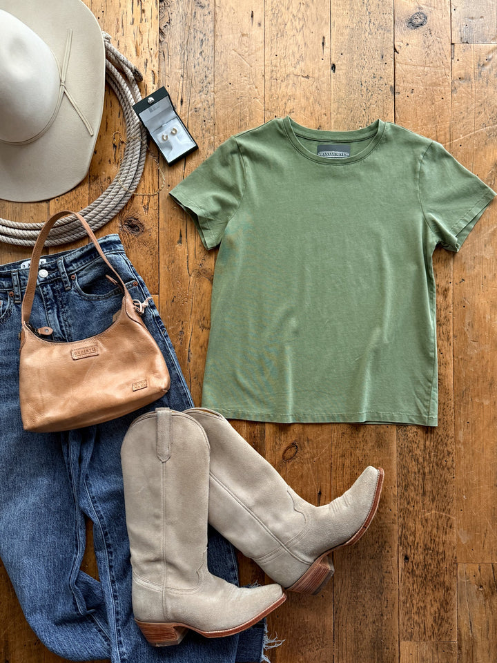 A Denning Moss Green Basic Tee laid out with blue jeans, beige cowboy boots, a tan leather shoulder bag, a wide-brimmed hat, a coiled rope, and a pair of small stud earrings.