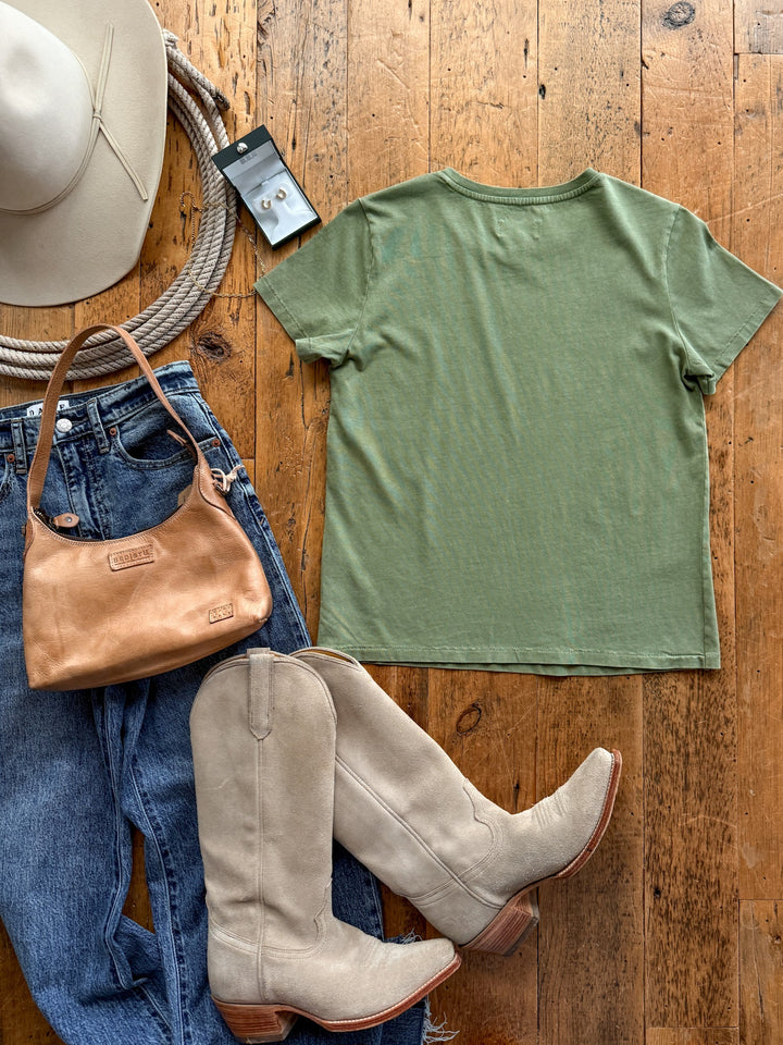 A moss green, relaxed-fit cotton Denning Moss Green Basic Tee laid out next to blue jeans, suede cowboy boots, a tan leather handbag, a wide-brimmed hat, a coiled rope, and a pair of stud earrings.