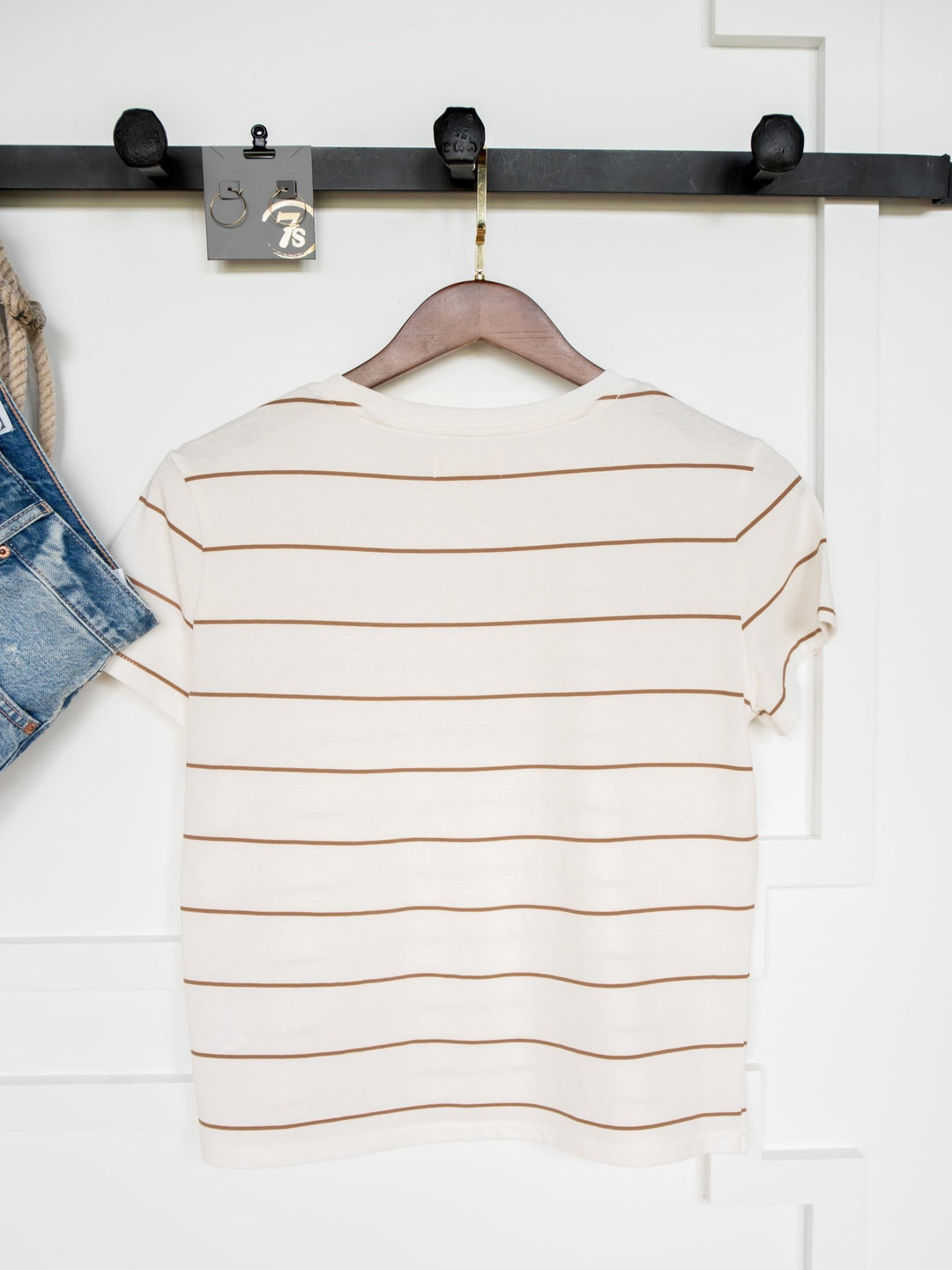 The Denning White Striped Tee by Savannah Sevens western life{&}style, a cream cotton short-sleeve with bronze stripes, hangs on a wooden hanger against a white background with denim shorts partially visible beside it.