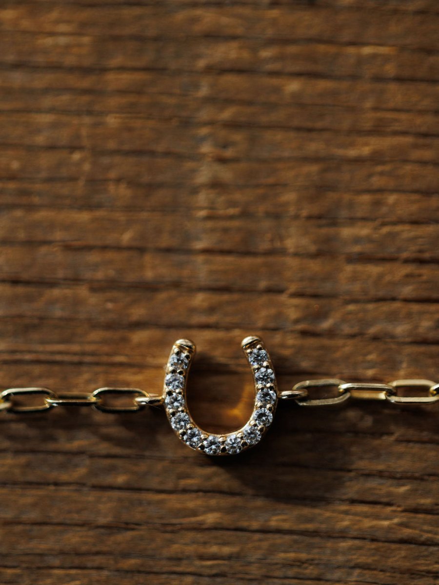 The Savannah Sevens western life{&}style Diamond Horseshoe Bracelet, featuring lab grown diamonds and 14K yellow gold links, rests on a textured wooden surface, its details illuminated by the light.