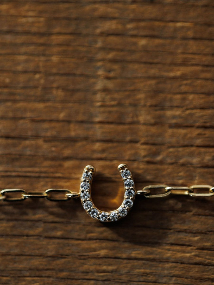 The Savannah Sevens western life{&}style Diamond Horseshoe Bracelet, featuring lab grown diamonds and 14K yellow gold links, rests on a textured wooden surface, its details illuminated by the light.