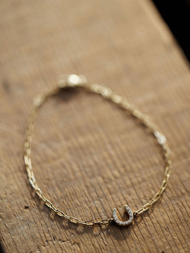 The Diamond Horseshoe Bracelet by Savannah Sevens western life{&}style features a delicate 14K yellow gold chain with a lab grown diamond-encrusted horseshoe charm, displayed on a wooden surface.