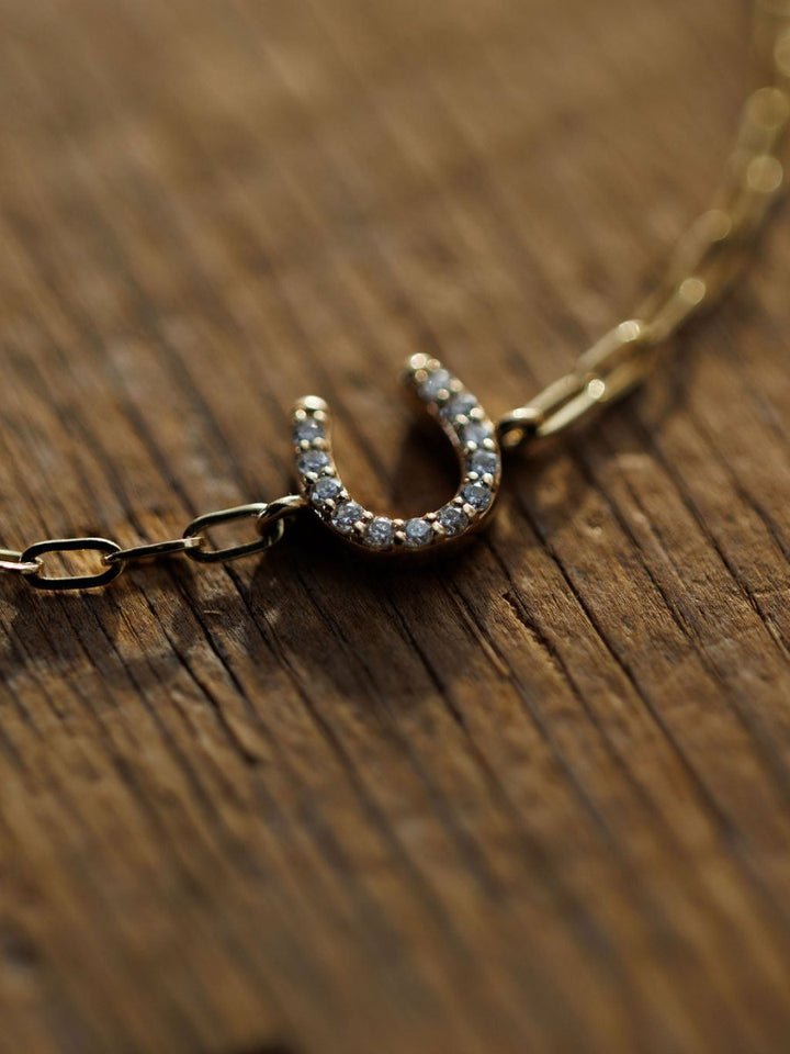 The Diamond Horseshoe Bracelet by Savannah Sevens western life{&}style, crafted in 14K yellow gold with a petite lab-grown diamond horseshoe charm, is displayed on a textured wooden surface.