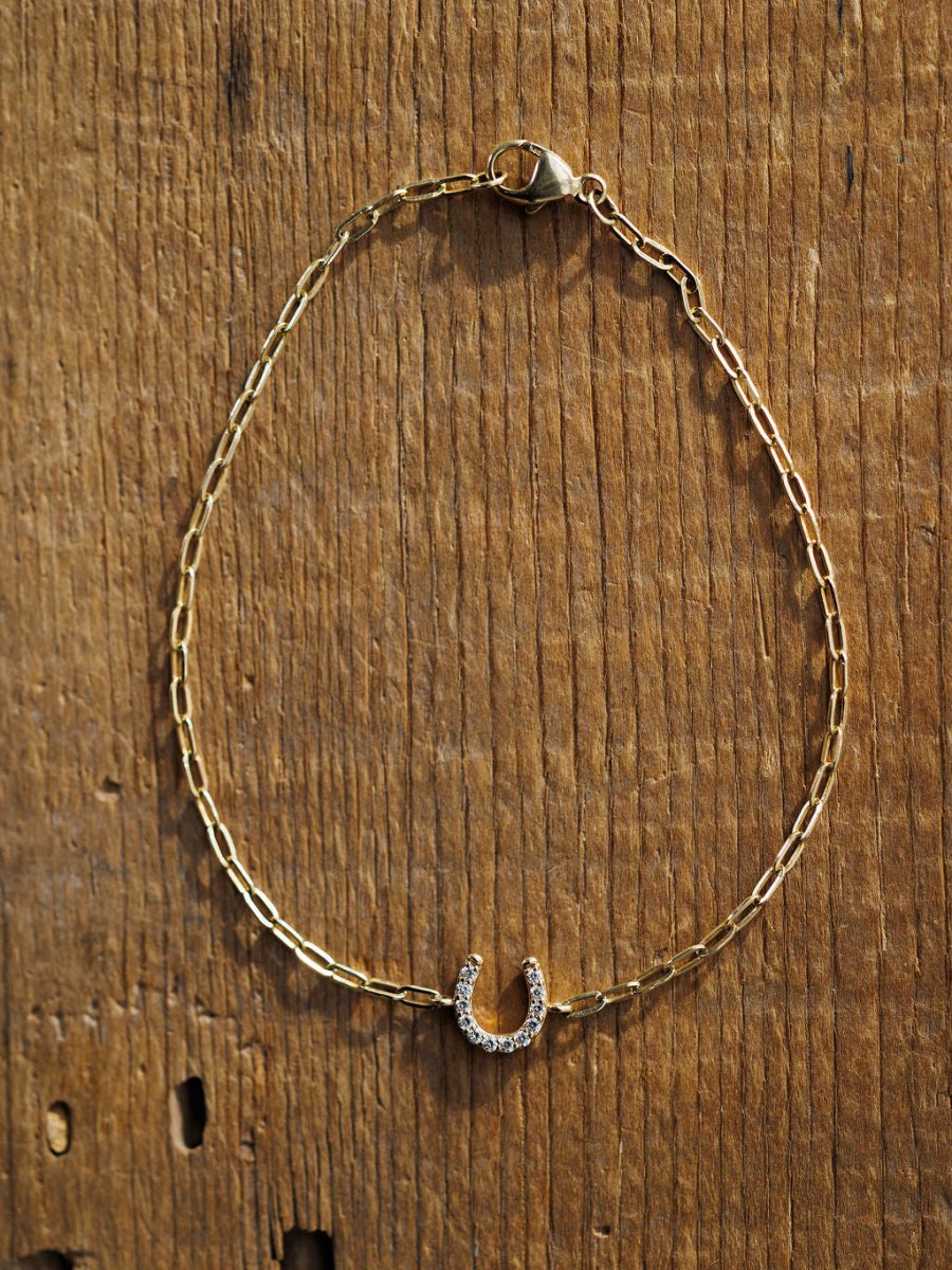 The Diamond Horseshoe Bracelet by Savannah Sevens western life{&}style is a 14K yellow gold piece featuring a lab grown diamond horseshoe charm, shown on a rustic wooden surface.