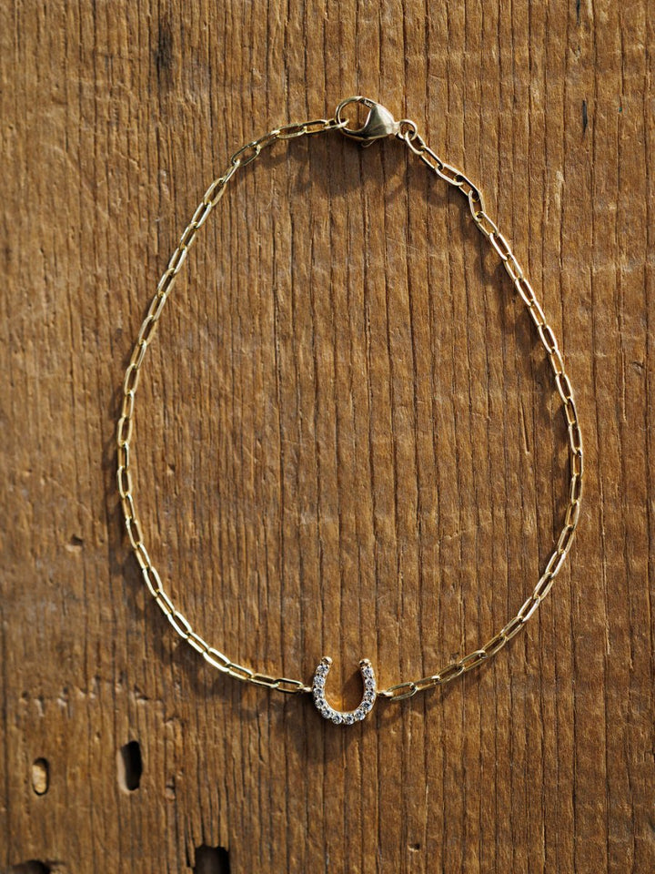 The Diamond Horseshoe Bracelet by Savannah Sevens western life{&}style is a 14K yellow gold piece featuring a lab grown diamond horseshoe charm, shown on a rustic wooden surface.