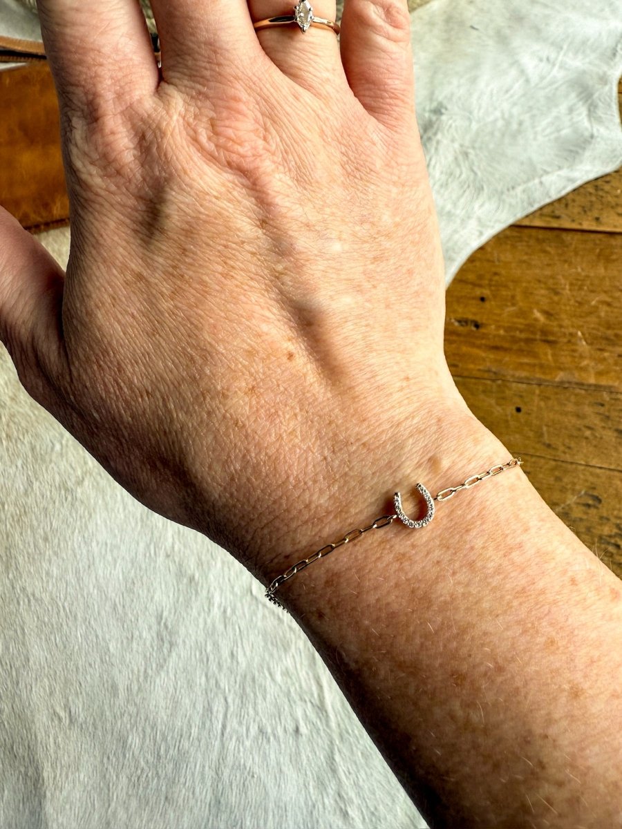 A hand wears the Savannah Sevens western life{&}style Diamond Horseshoe Bracelet in 14K yellow gold with a small diamond horseshoe charm, alongside a gold ring with a clear stone, resting on a light surface.