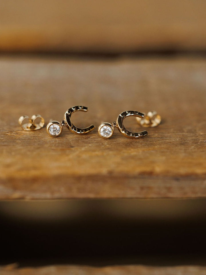 The Diamond Horseshoe Drop Earrings by Savannah Sevens western life{&}style are 14K yellow gold with a lab grown diamond at each opening, shown on a wooden surface.