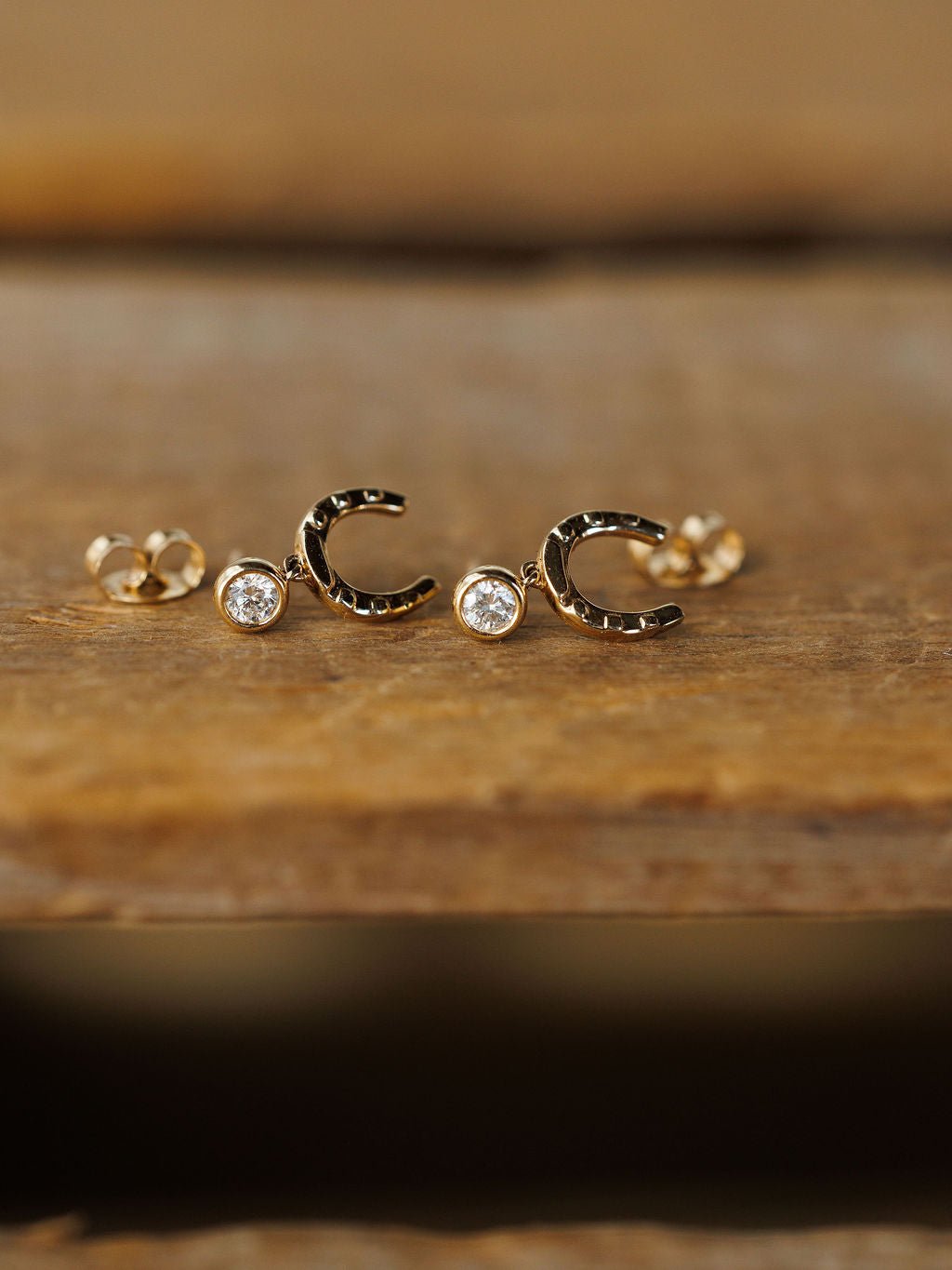 The Diamond Horseshoe Drop Earrings by Savannah Sevens western life{&}style are 14K yellow gold with a lab grown diamond at each opening, shown on a wooden surface.