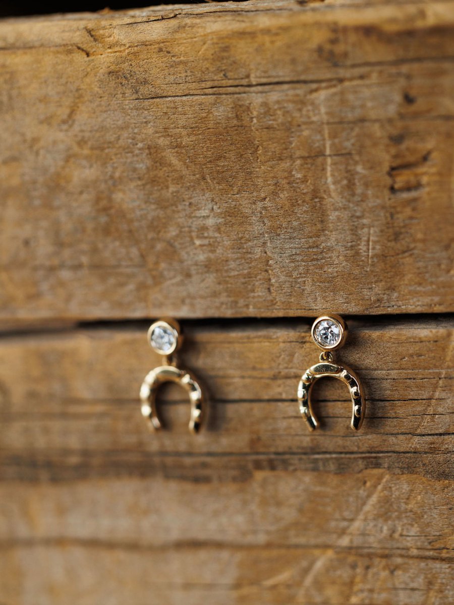 The Diamond Horseshoe Drop Earrings by Savannah Sevens western life{&}style feature 14K yellow gold with round lab grown diamonds atop diamond horseshoe charms, shown on a rustic wooden surface.