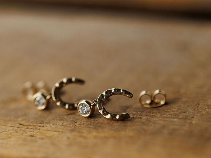 The Diamond Horseshoe Drop Earrings by Savannah Sevens western life{&}style feature 14K yellow gold horseshoe shapes with small round lab grown diamonds, stylishly displayed on a wooden surface.