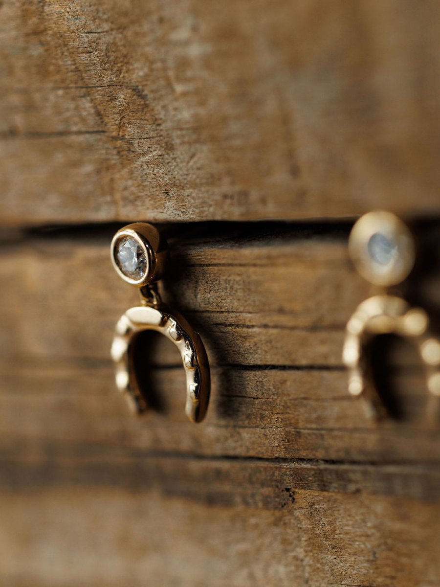 Close-up of Savannah Sevens western life{&}style Diamond Horseshoe Drop Earrings in 14K yellow gold with small lab-grown diamonds, displayed on a rustic wooden surface with soft focus emphasizing the wood texture and earring details.