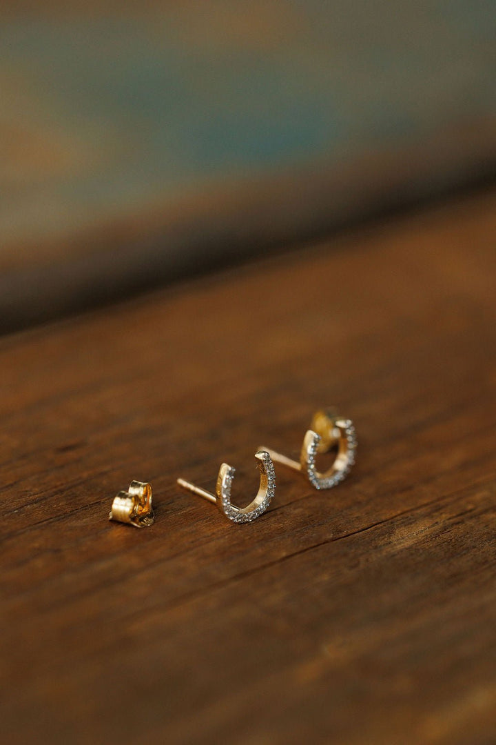 The Diamond Horseshoe Earrings by Savannah Sevens western life{&}style, crafted in 14K gold and adorned with sparkling clear stones, rest on a rustic wooden surface.