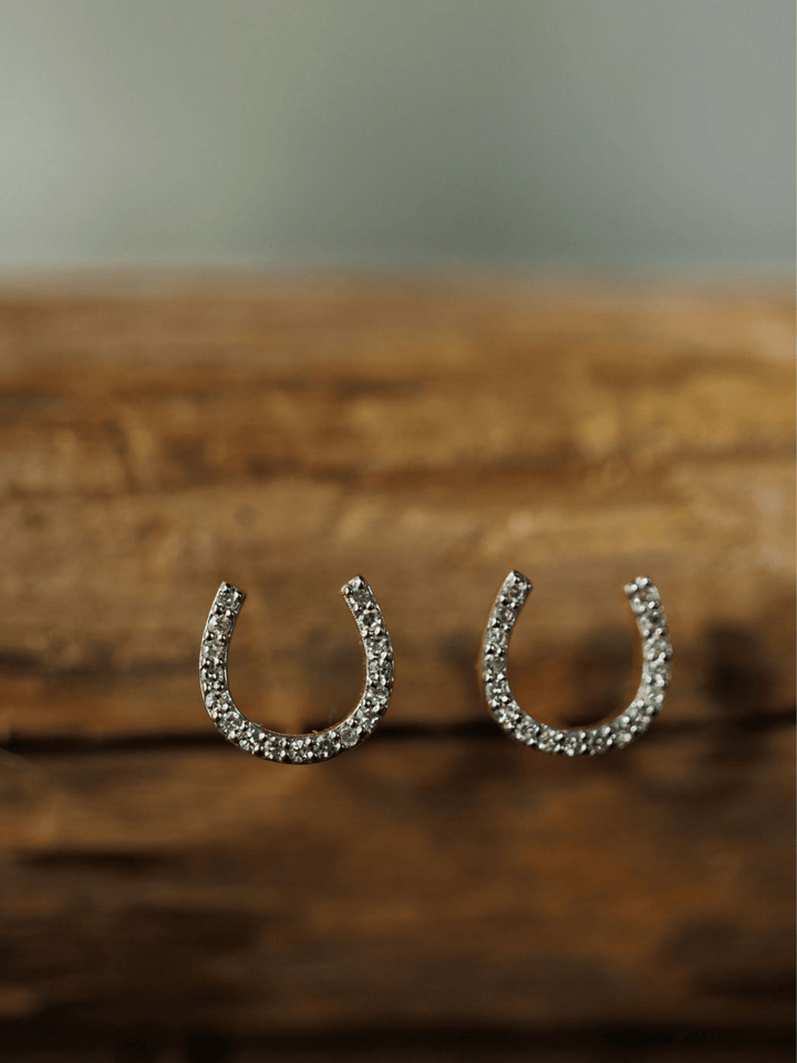 The Diamond Horseshoe Earrings by Savannah Sevens western life{&}style, encrusted with sparkling stones, are showcased against a rustic wooden background.