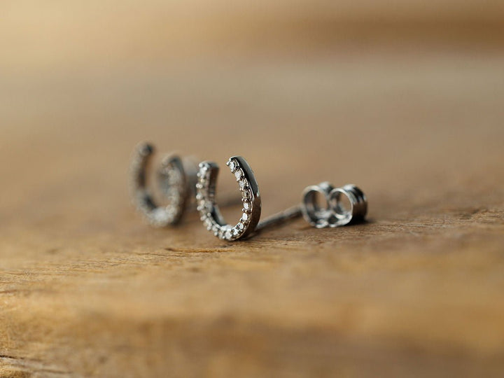 The Diamond Horseshoe Earrings by Savannah Sevens western life{&}style are small, silver earrings featuring embedded clear stones, shown on a wooden surface.