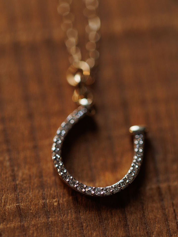 The Savannah Sevens western life{&}style Diamond Horseshoe Necklace, featuring sparkling clear stones on a delicate 14K gold chain, is displayed elegantly on a wooden surface.