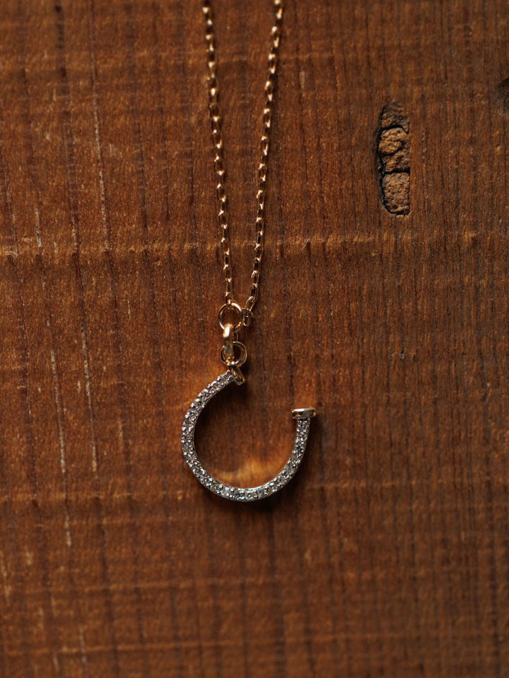 The Diamond Horseshoe Necklace by Savannah Sevens western life{&}style features a sparkling horseshoe pendant, shown on a wooden surface.