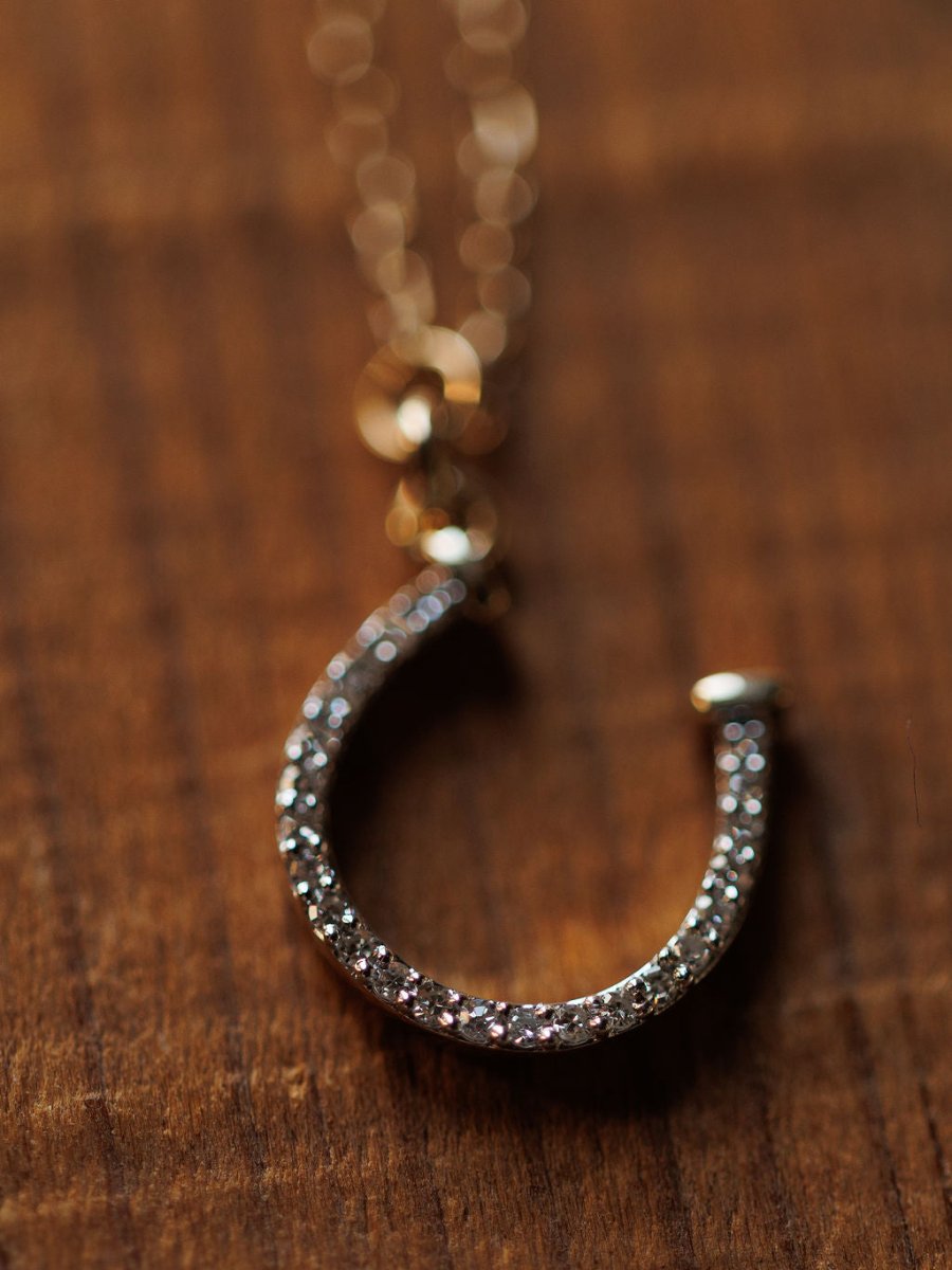 The Savannah Sevens western life{&}style Diamond Horseshoe Necklace, featuring sparkling clear stones on a delicate 14K gold chain, is displayed elegantly on a wooden surface.