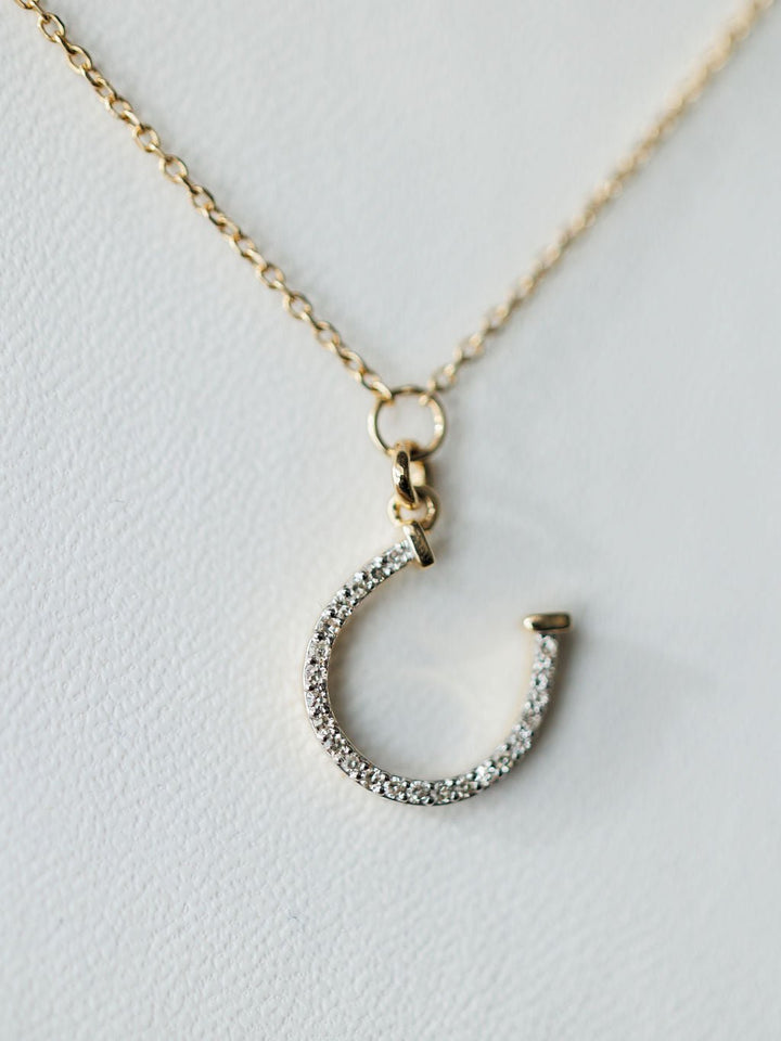 A close-up of the Diamond Horseshoe Necklace by Savannah Sevens western life{&}style, a 14K gold chain with a diamond-encrusted horseshoe pendant, photographed against a white background.
