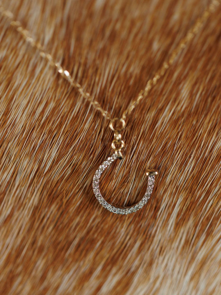 The Savannah Sevens western life{&}style Diamond Horseshoe Necklace in 14K gold is displayed on a textured brown and white fur background.
