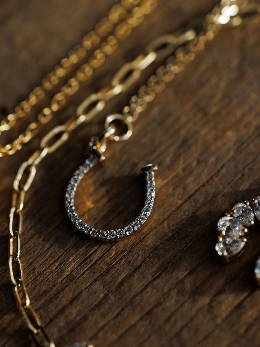 A close-up of Savannah Sevens western life{&}style's Diamond Horseshoe Necklace, 14K gold with sparkling diamonds, rests on wood beside gold chains and a jeweled accessory.  Devereaux Gold.