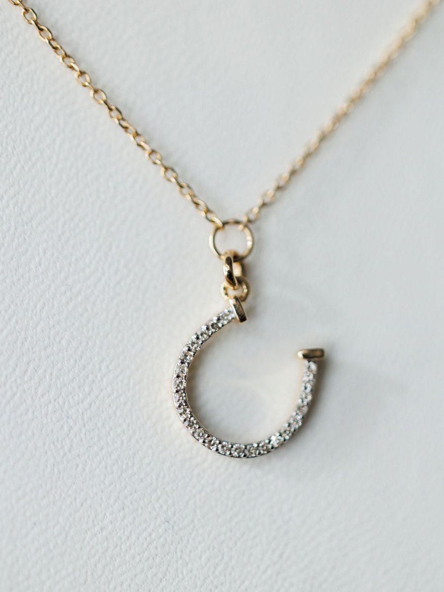 A close-up of the Diamond Horseshoe Necklace by Savannah Sevens western life{&}style, a 14K gold chain with a diamond-encrusted horseshoe pendant, photographed against a white background.