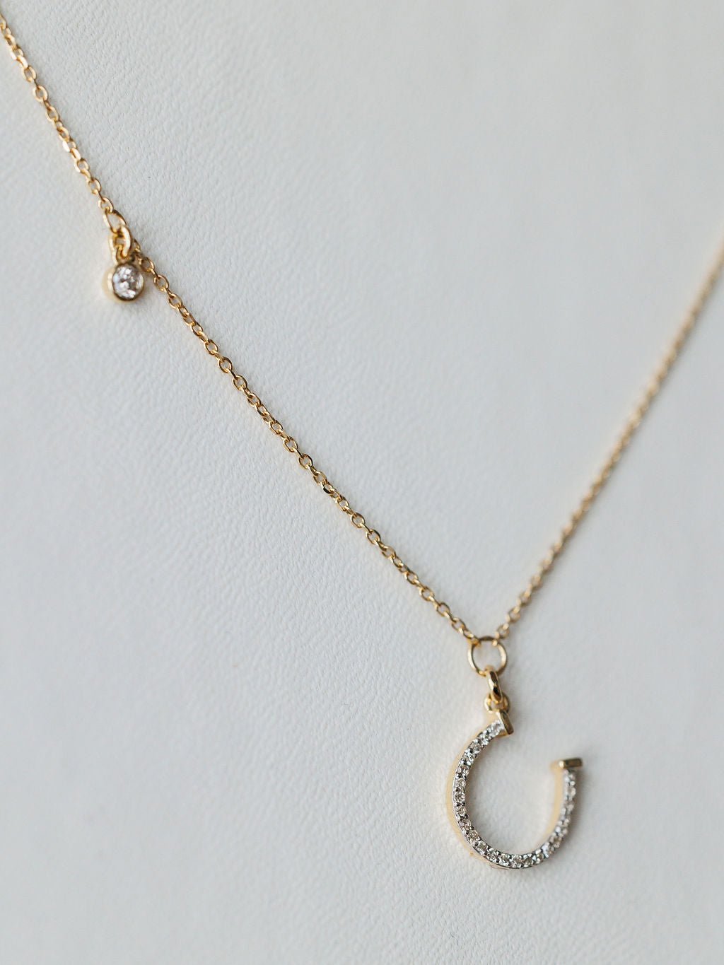 The Savannah Sevens western life{&}style Diamond Horseshoe Necklace features a delicate gold chain with a small round diamond charm and a 14K gold horseshoe pendant encrusted with sparkling stones, shown on a light background.